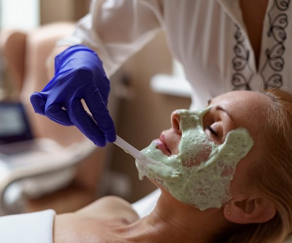 Hands of cosmetology specialist applying facial alginate mask using wooden spatula, making skin hydrated and face glowing and skin.